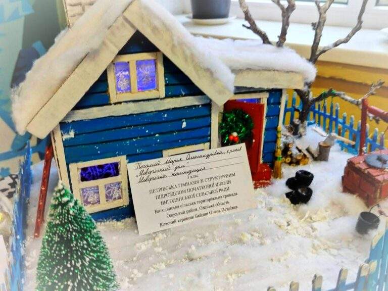 Мініатюрна зимова сцена: синій дерев’яний будиночок зі сніжним дахом, червоними дверима з вінком, білим оздобленням вікон і засніженим подвір’ям. У дворі — синій паркан, ялинка з прикрасами, криниця, дрова, мітла, чорні горщики та засніжене дерево. Перед будинком — інформаційна картка з даними про автора композиції, ученицю 6 класу Плохій Марію Олександрівну, та її школу в Одеській області.